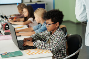 Side view of youthful African American schoolboy and his classmates working in front of laptops while sitting in row by desk