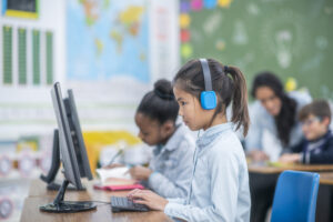 A diverse group of girls in a primary school class work on computers, wearing headphones.